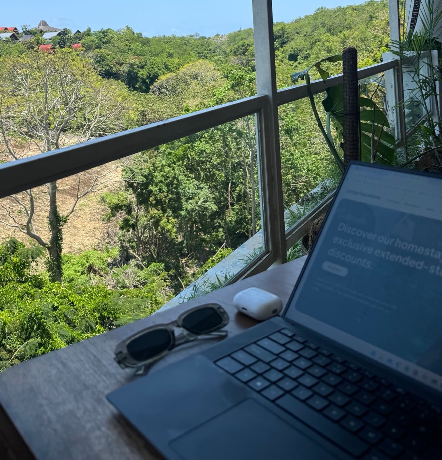 A laptop open to the Lodge Vacations website on a jungle-view balcony in Bali, with sunglasses and wireless earbuds on the table.